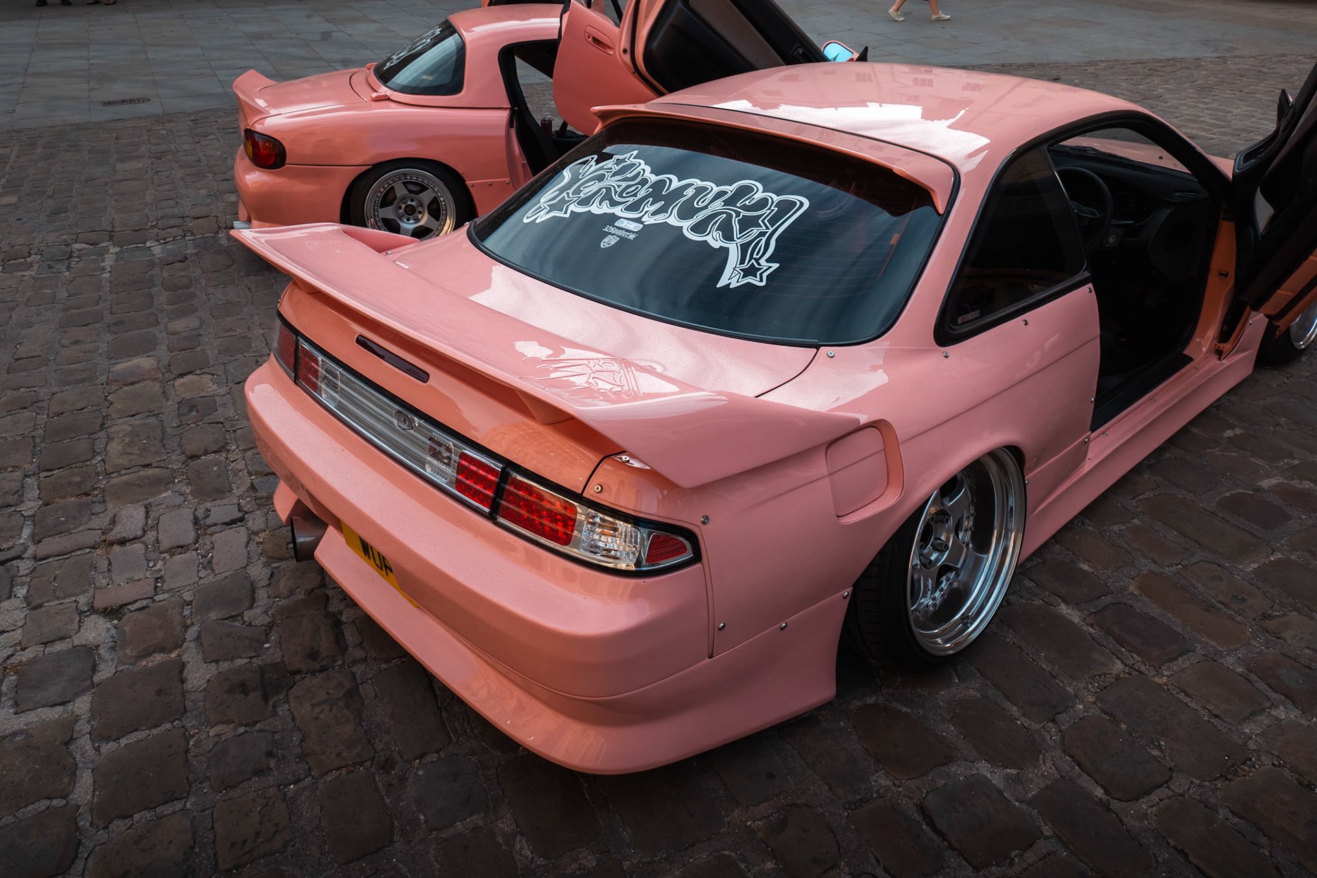 Professional rear quarter shot of two modified Japanese cars in a soft pink colour, parked side-by-side with suicide doors raised up.